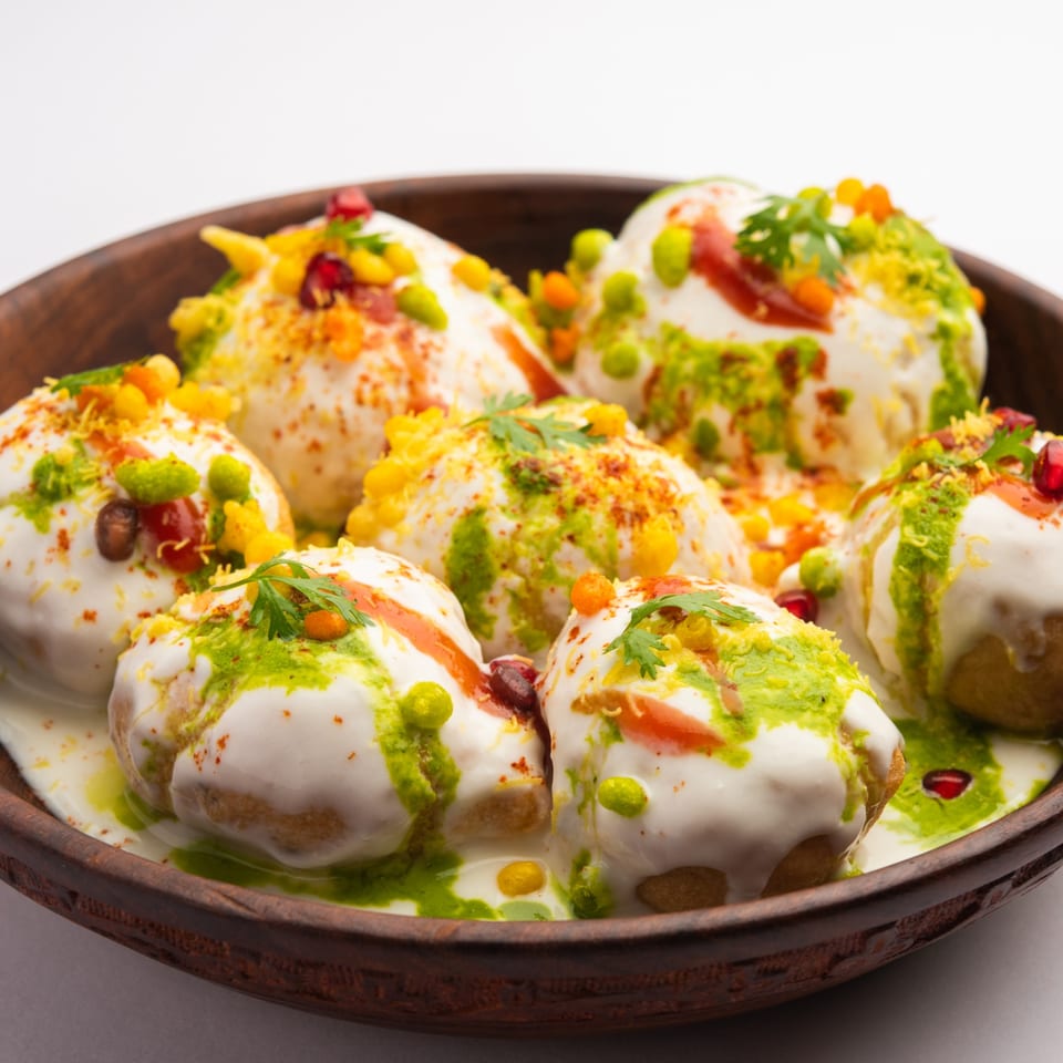 A wooden bowl filled with Bombay Chaat House's Dahi Vada, topped with yogurt, chutneys, and spices.