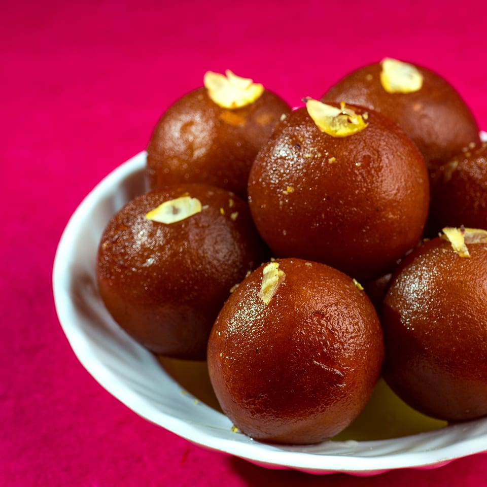 A white bowl filled with glistening Gulab Jamun, garnished with slivered almonds, against a vibrant pink background.