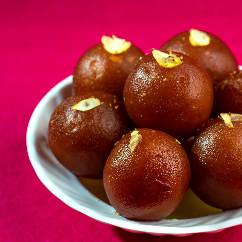 A white bowl filled with glistening Gulab Jamun, garnished with slivered almonds, against a vibrant pink background.