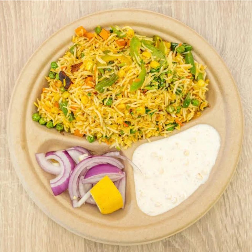 Vegetable biryani with red onions, a lemon wedge, and raita on a divided plate.