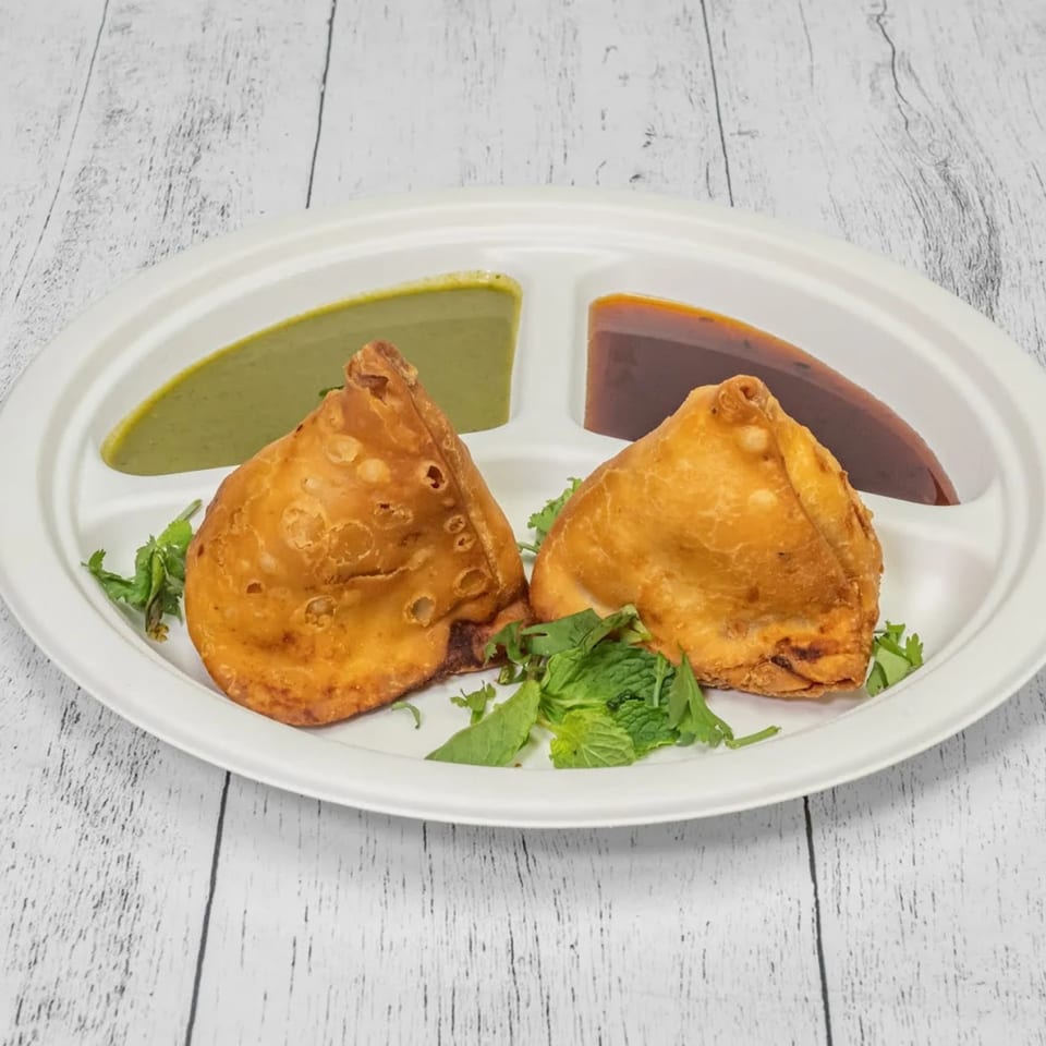 Two golden-brown samosas with mint leaves, green chutney, and tamarind chutney on a white plate.