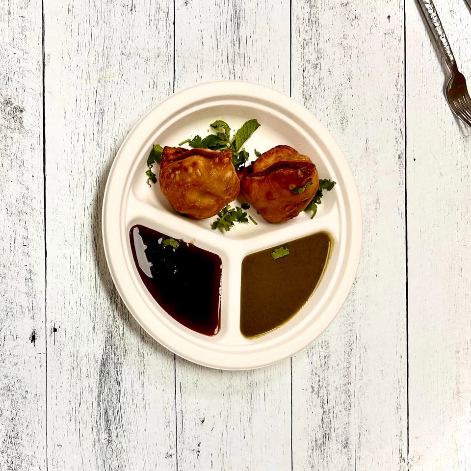 Two golden-brown samosas with cilantro on a divided plate, served with dark and green chutneys.